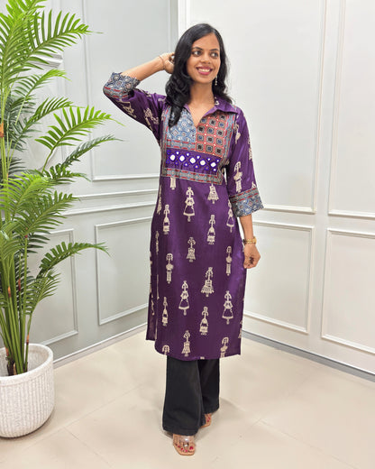 Purple women's Pure Mashru Silk Long Kurti with red , Black & white geometric patterns on the yoke and sleeves, paired with black Flared pants, standing in a room with a potted plant.
