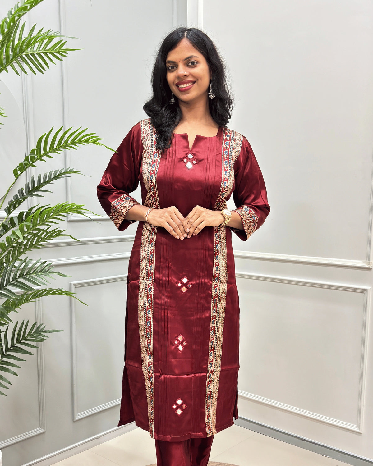 Front View of Deep Red women's Ajrakh Mashru Silk with beige and multicolor embroidered borders, paired with matching pant , standing in front of a white paneled wall with a green plant on the left.