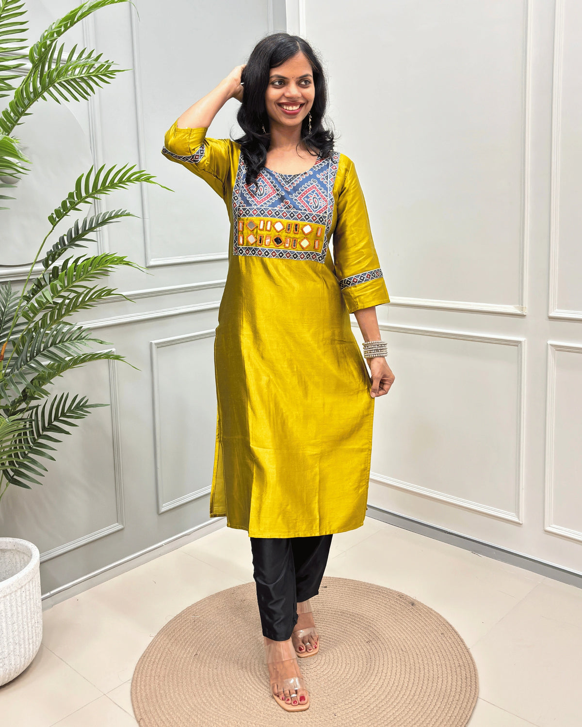 Mustard Yellow Women's Ajrakh Mahru Silk Long-kurti with a geometric Print yoke in blue, red, and white, with Hand-Mirror Work,  paired with black pants, standing on a round woven mat in front of a white paneled wall with a potted plant.