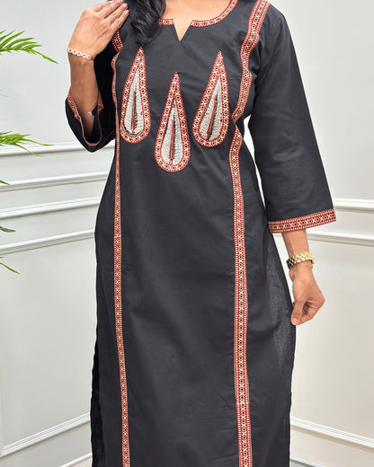 Close-up of Black Women's Hand-block  Ajrakh Pure Cotton Long-kurti with three-quarter sleeve featured with intricate Beige, Black, and Red embroidered details on the neckline, cuffs, and front  panel, Along with Grey on  Palm Leaf set against a light gray wall with a plant in the background.