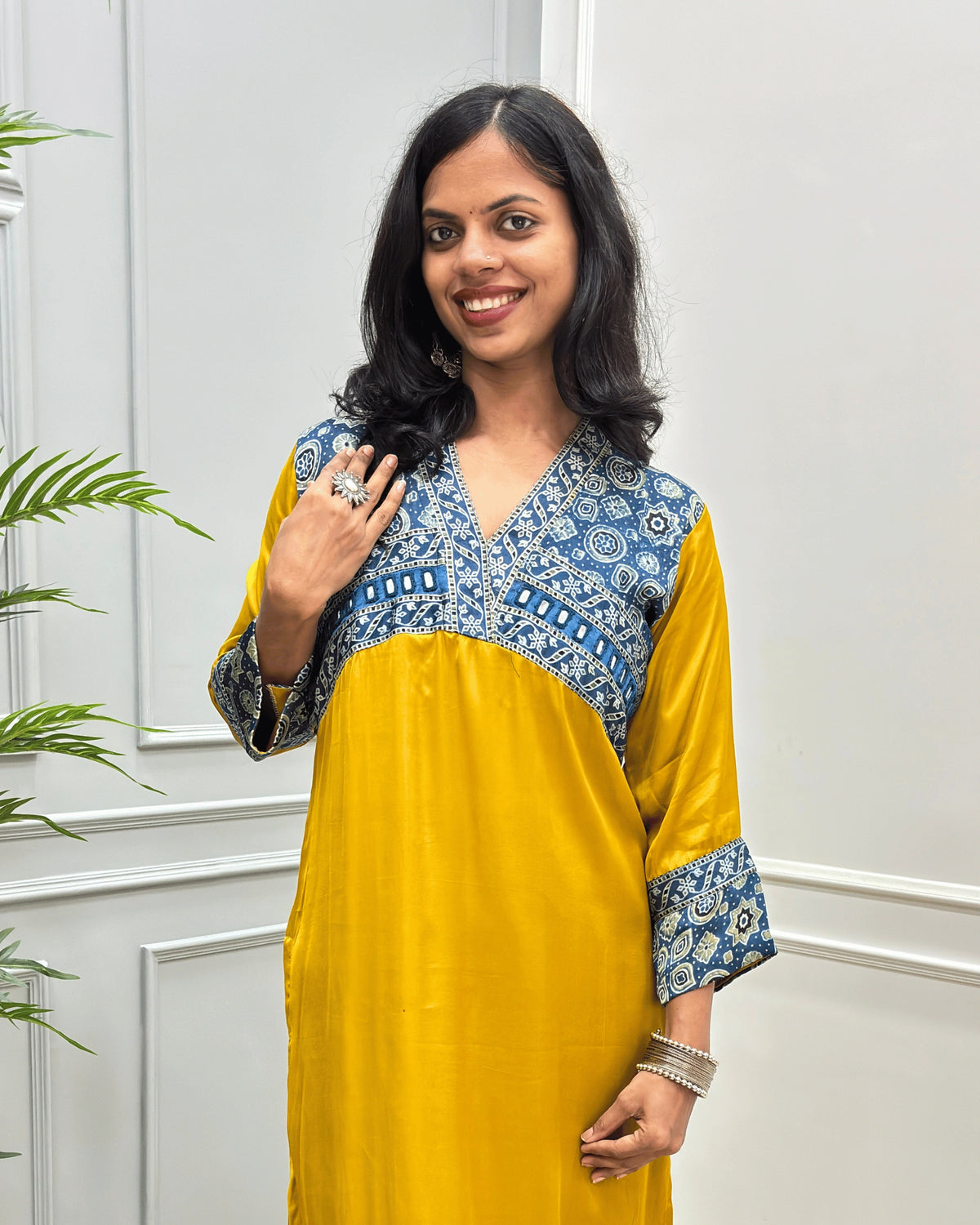 Close-up of Mustard-Yellow Ajrakh Model silk fabric with three-quarter sleeves with intricate blue and white patterned embroidery on the V-neckline, chest panel, and cuffs, standing in a light-colored room. 