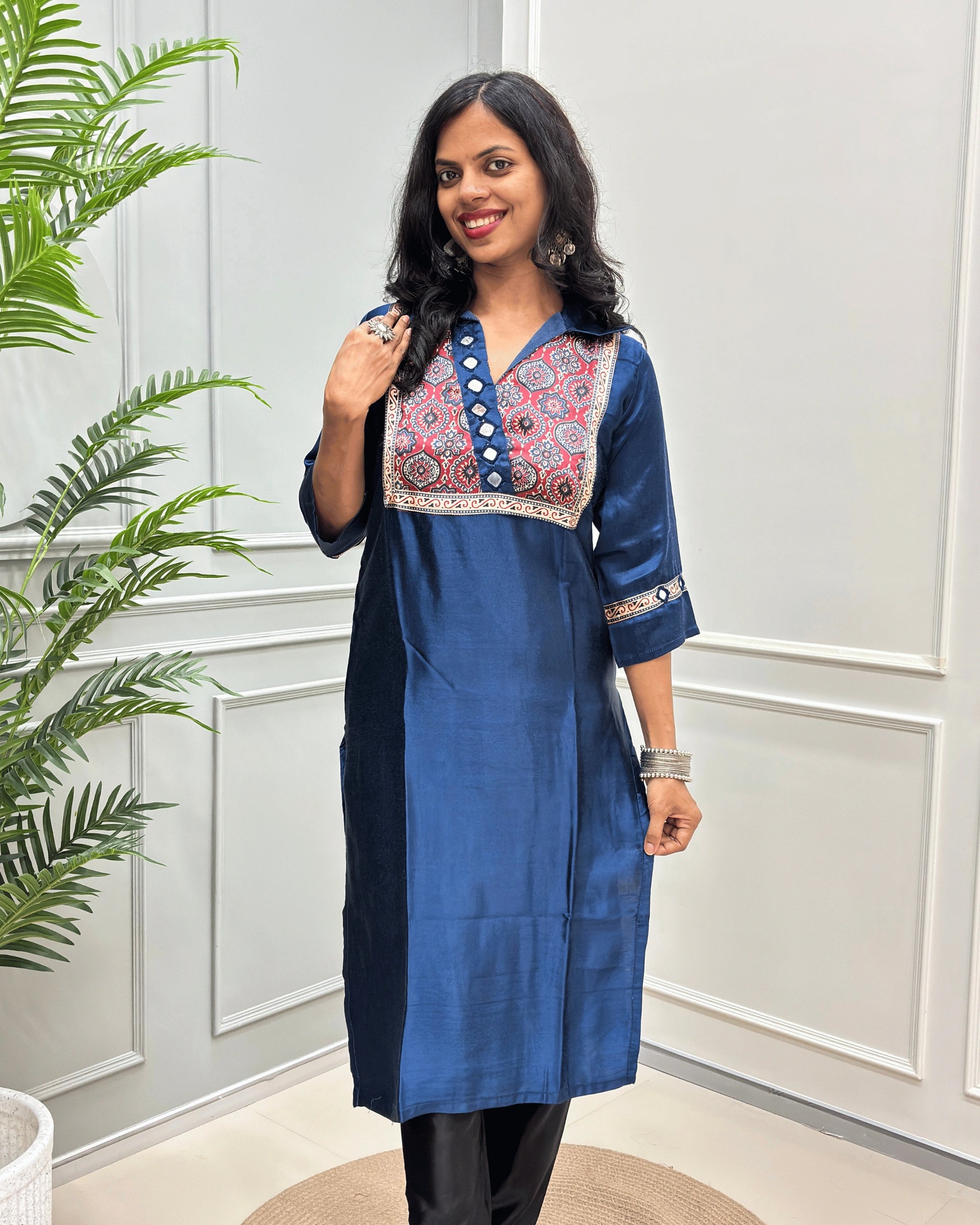 Front view of Blue women's Ajrakh Mashru Silk with three-quarter Sleeves Featured with detailed multicolor embroidered yoke and patterned sleeve cuffs with Badam Buta print , standing in front of a white paneled wall with a green plant on the left.