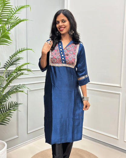 Front view of Blue women's Ajrakh Mashru Silk with three-quarter Sleeves Featured with detailed multicolor embroidered yoke and patterned sleeve cuffs with Badam Buta print , standing in front of a white paneled wall with a green plant on the left.