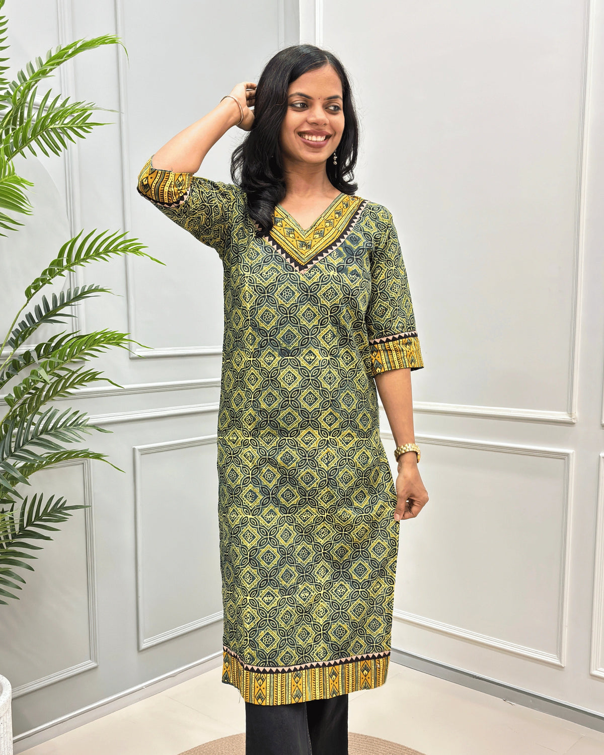Front View of Green Women's Ajrakh Hand-block Cotton Long Kurti with , Green, yellow, and Black ajrakh Print on V-neck ,Strip print on trim & Sleeves , standing in front of a white paneled wall with a green plant on the left.