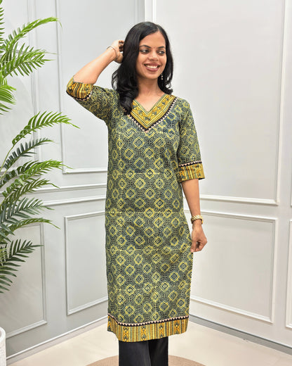 Front View of Green Women's Ajrakh Hand-block Cotton Long Kurti with , Green, yellow, and Black ajrakh Print on V-neck ,Strip print on trim & Sleeves , standing in front of a white paneled wall with a green plant on the left.