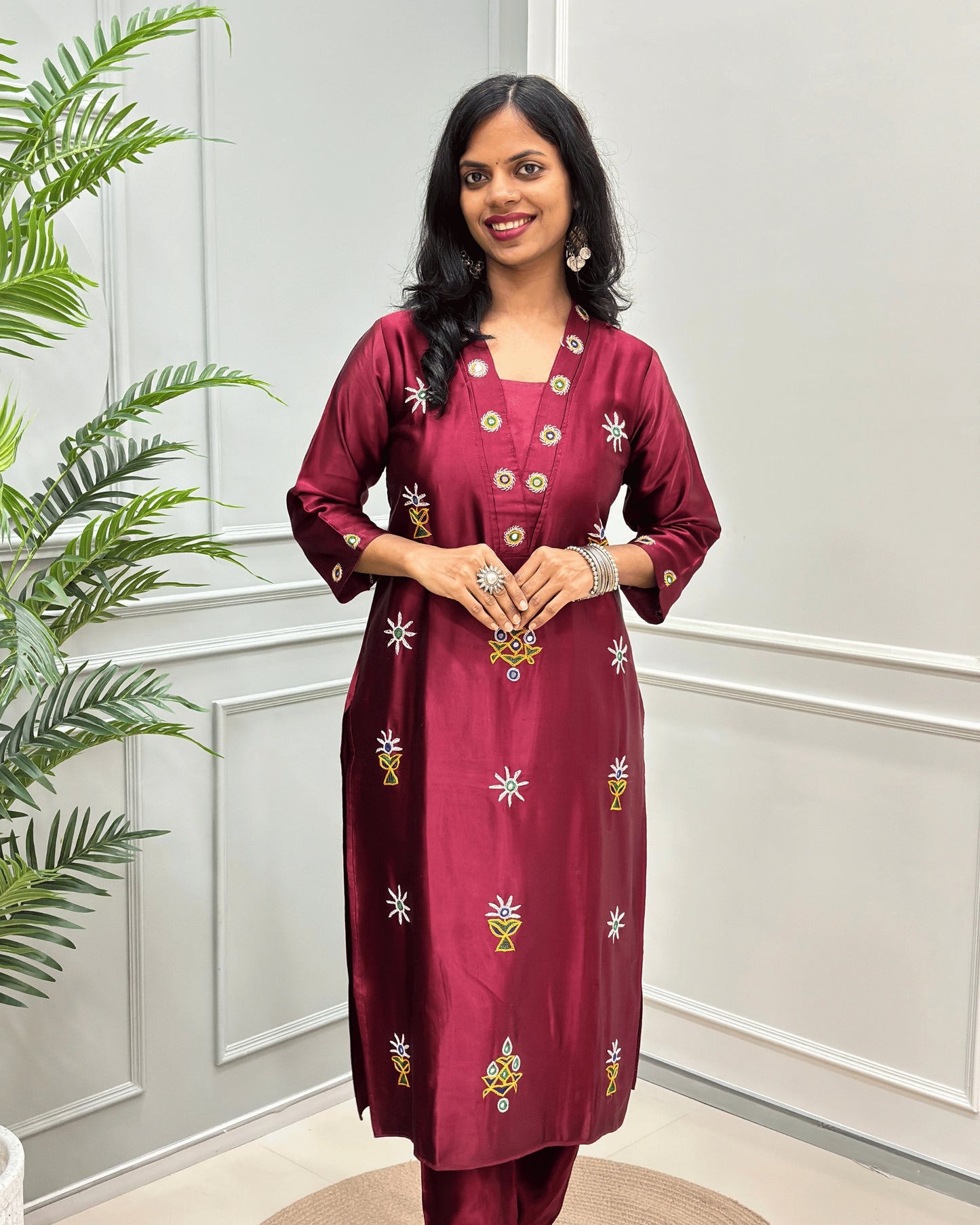 Front view of Maroon women's pure Mashru Silk with embroidered Ahir motifs, Hand-mirror work on front, V shaped neck, & Sleeves cuff,  standing in front of a white paneled wall with a green plant on the left.