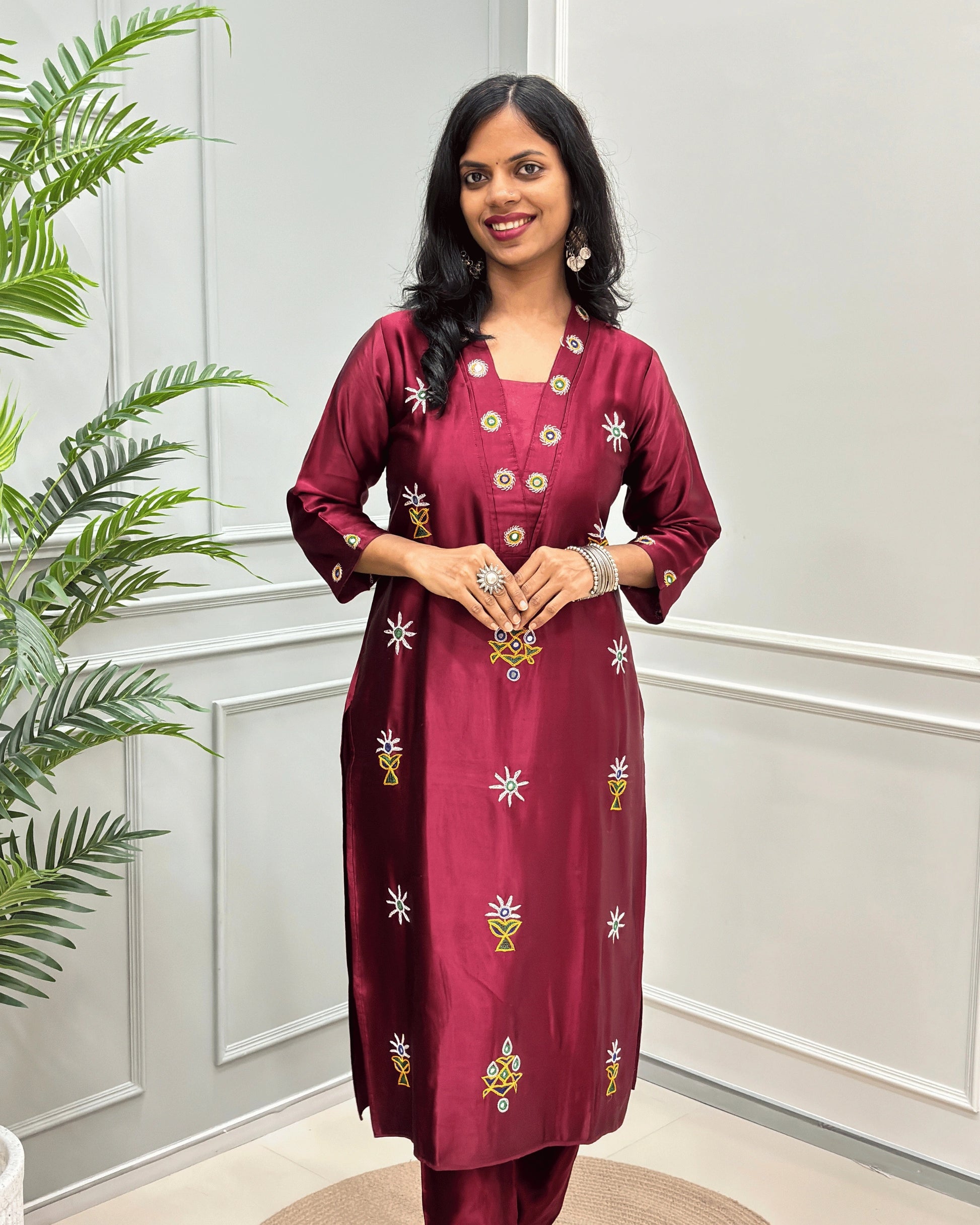 Front view of Maroon women's pure Mashru Silk with embroidered Ahir motifs, Hand-mirror work on front, V shaped neck, & Sleeves cuff,  standing in front of a white paneled wall with a green plant on the left.