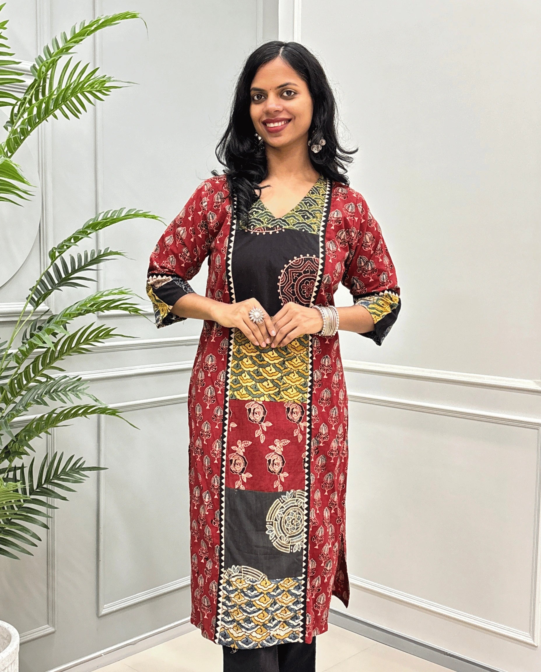 Front view of Red women's Hand-block Ajrakh pure cotton long kurti with three quarter full printed sleeves with a Red base, & Abstract red, yellow, and black Ajrakh Geometric print panel & cuffs, standing in front of a light gray paneled wall with a green plant on the left.