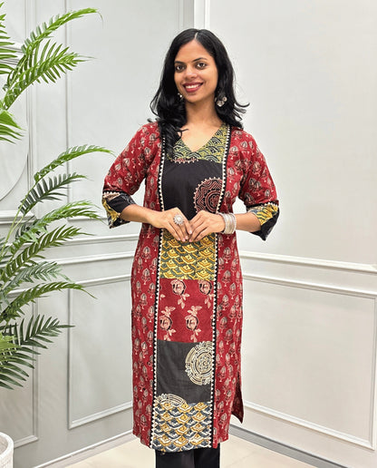 Front view of Red women's Hand-block Ajrakh pure cotton long kurti with three quarter full printed sleeves with a Red base, & Abstract red, yellow, and black Ajrakh Geometric print panel & cuffs, standing in front of a light gray paneled wall with a green plant on the left.