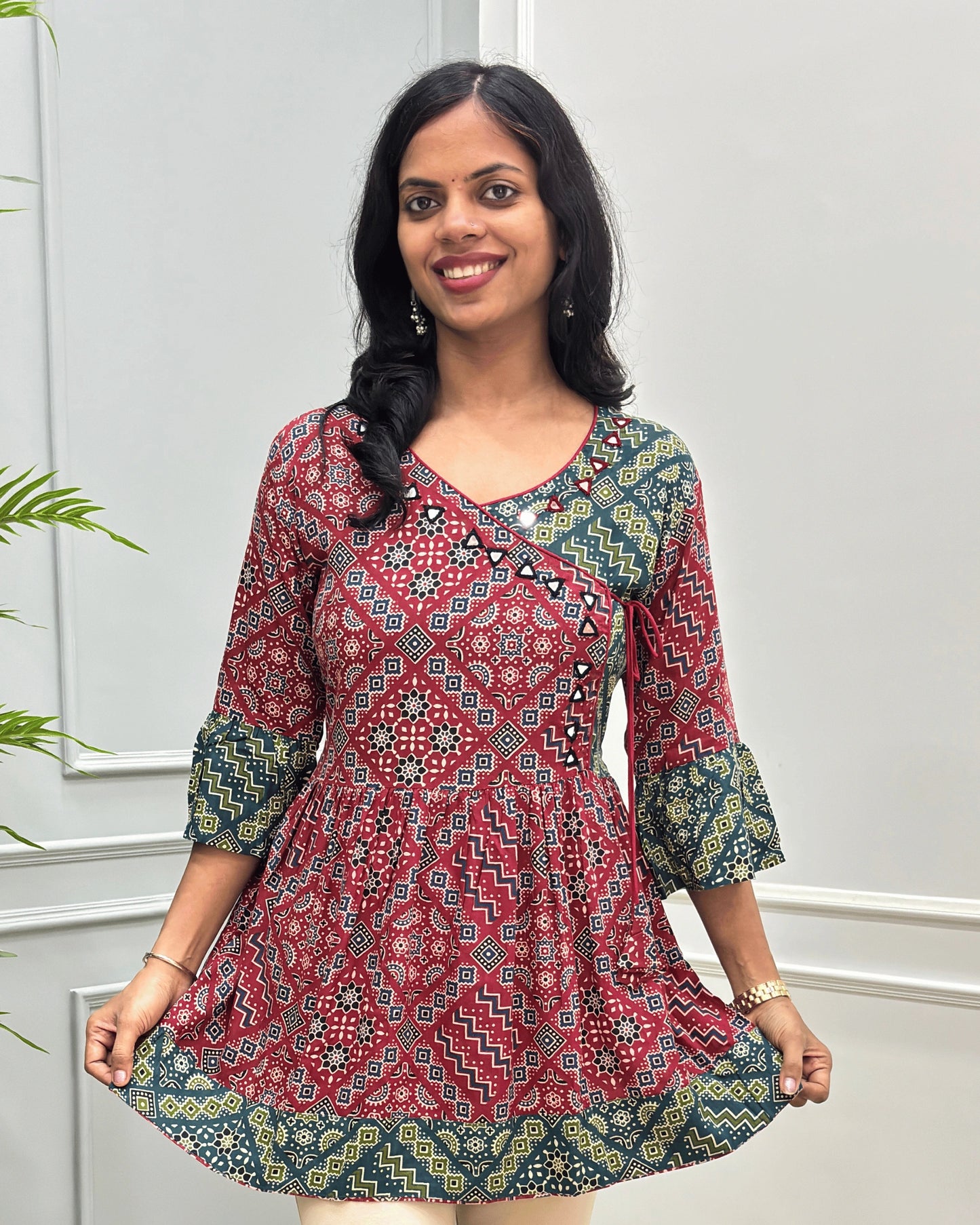 Close-up of Red women's Ajrakh Plant based cotton short-kurti with a Three-Quarter Sleeves (frill)  featuring Red & Green Geometric print ,  Angrakha Pattern with Hand-mirror work on V-shaped Neckline along with Lace, set against a light gray paneled wall.