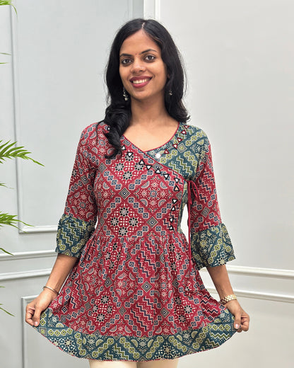 Close-up of Red women's Ajrakh Plant based cotton short-kurti with a Three-Quarter Sleeves (frill)  featuring Red & Green Geometric print ,  Angrakha Pattern with Hand-mirror work on V-shaped Neckline along with Lace, set against a light gray paneled wall.