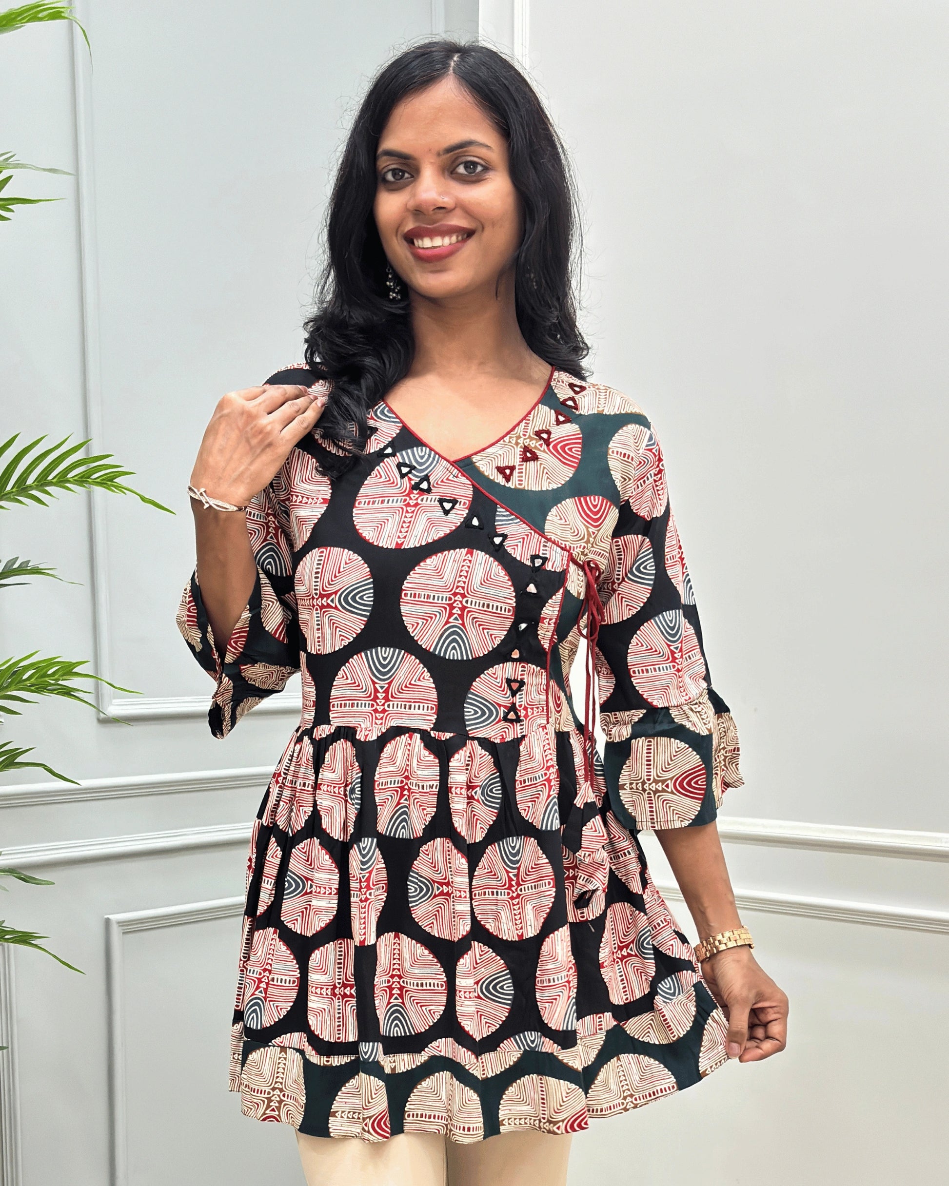 Close-up of Black women's Ajrakh plant based cotton Short kurti with a colorful geometric and circular print, Hand-Mirror work on V- neckline along with lace, paired with beige pants, standing in front of a white paneled wall with a green plant on the left.