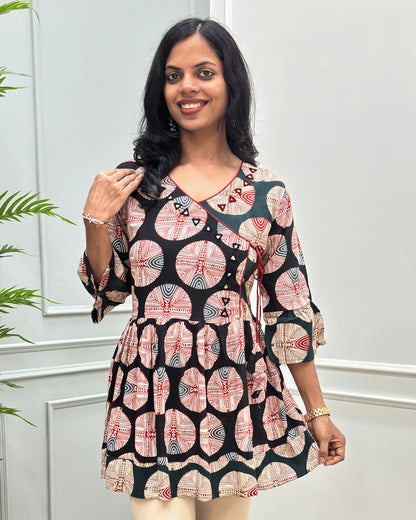 Close-up of Black women's Ajrakh plant based cotton Short kurti with a colorful geometric and circular print, Hand-Mirror work on V- neckline along with lace, paired with beige pants, standing in front of a white paneled wall with a green plant on the left.