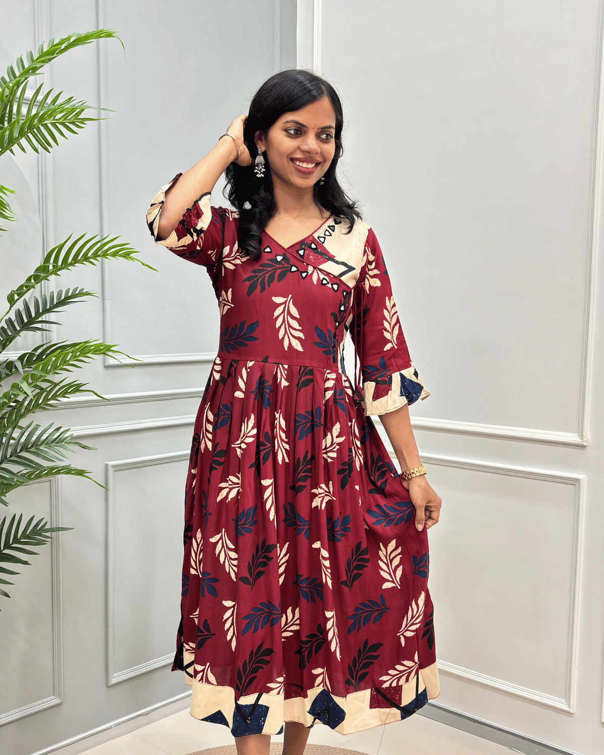 Front view of Red Women's Plant based cotton long kurti with a navy and beige leaf print along with Angrakha pattern , standing in front of a white paneled wall with a green plant on the left.