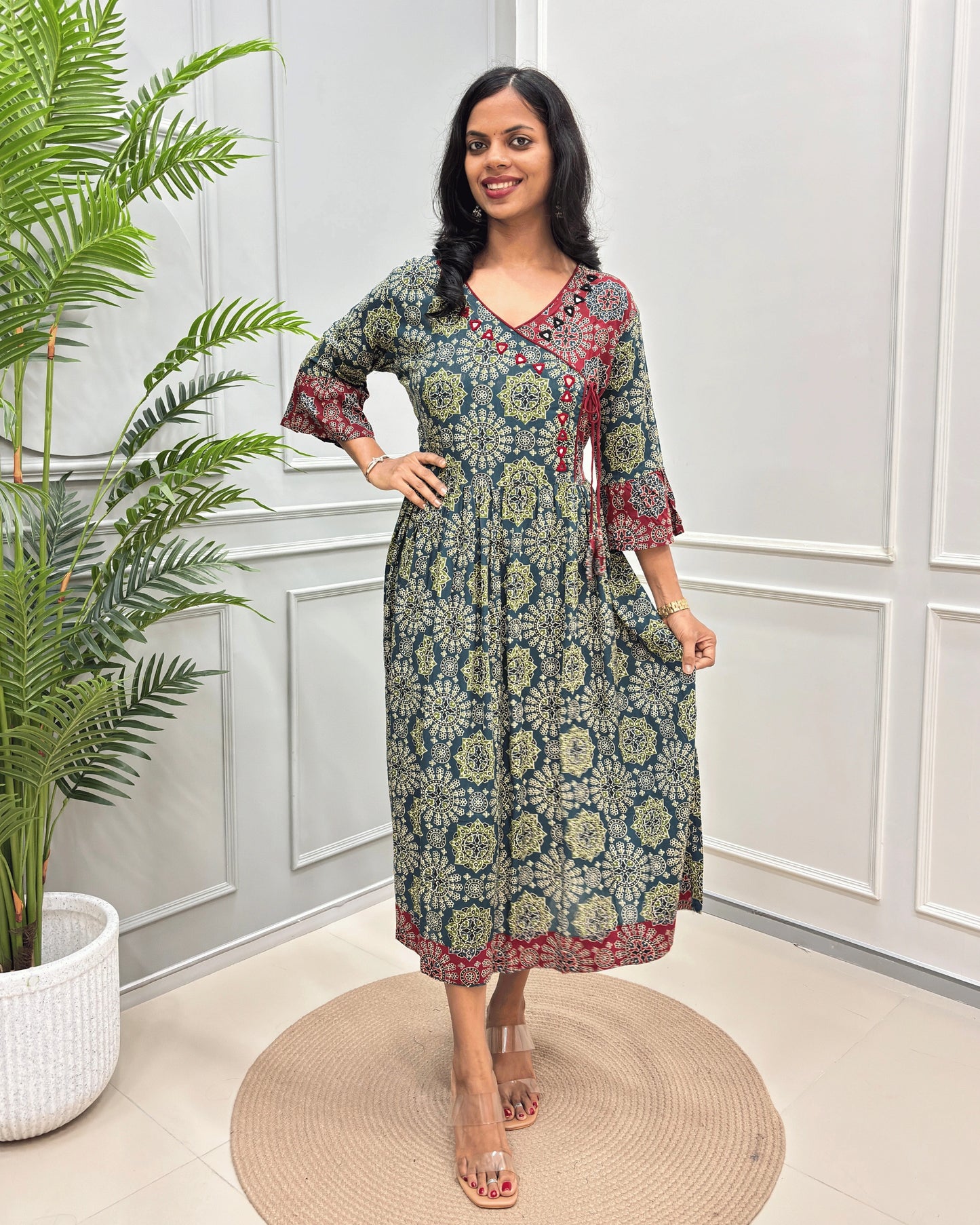 Green-red woman's Cotton long-kurti with Hand-mirror work on V-neck, a green, beige, and red floral mandala pattern,  standing in a room with white paneled walls and a potted plant.