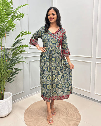 Green-red woman's Cotton long-kurti with Hand-mirror work on V-neck, a green, beige, and red floral mandala pattern,  standing in a room with white paneled walls and a potted plant.